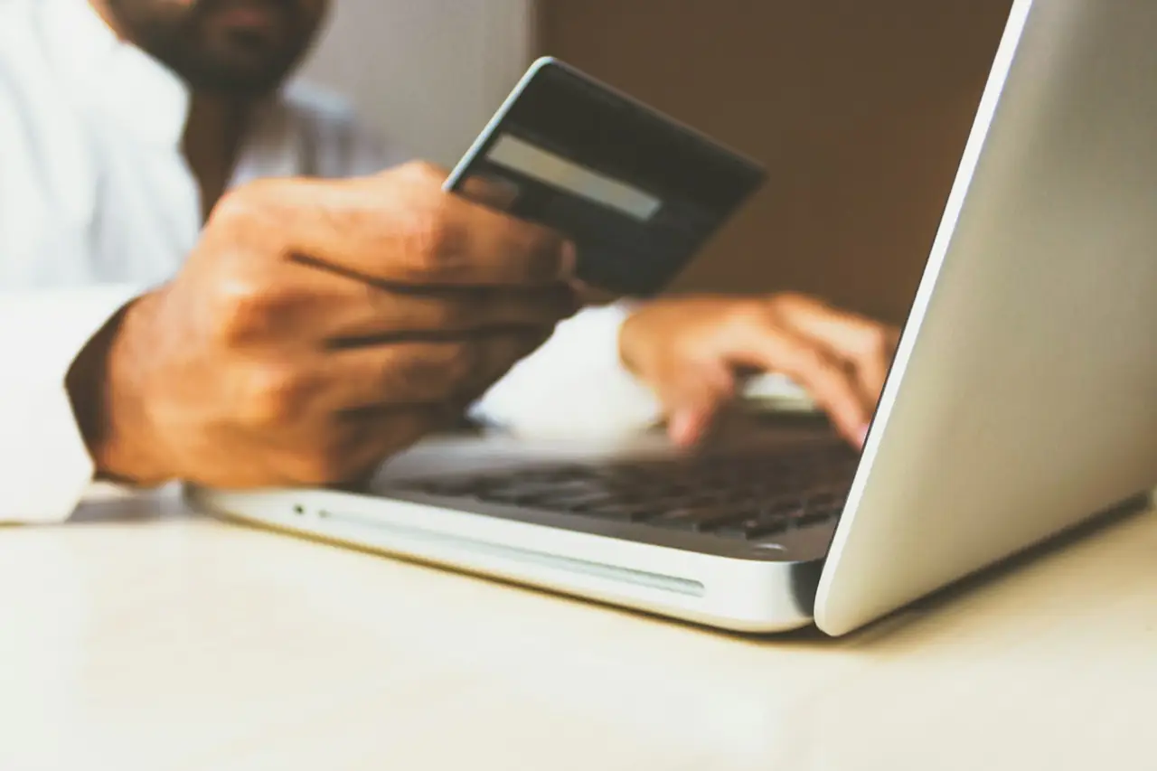 Persoană care ține un card bancar în mână în fața unui laptop deschis, pregătind o plată online.